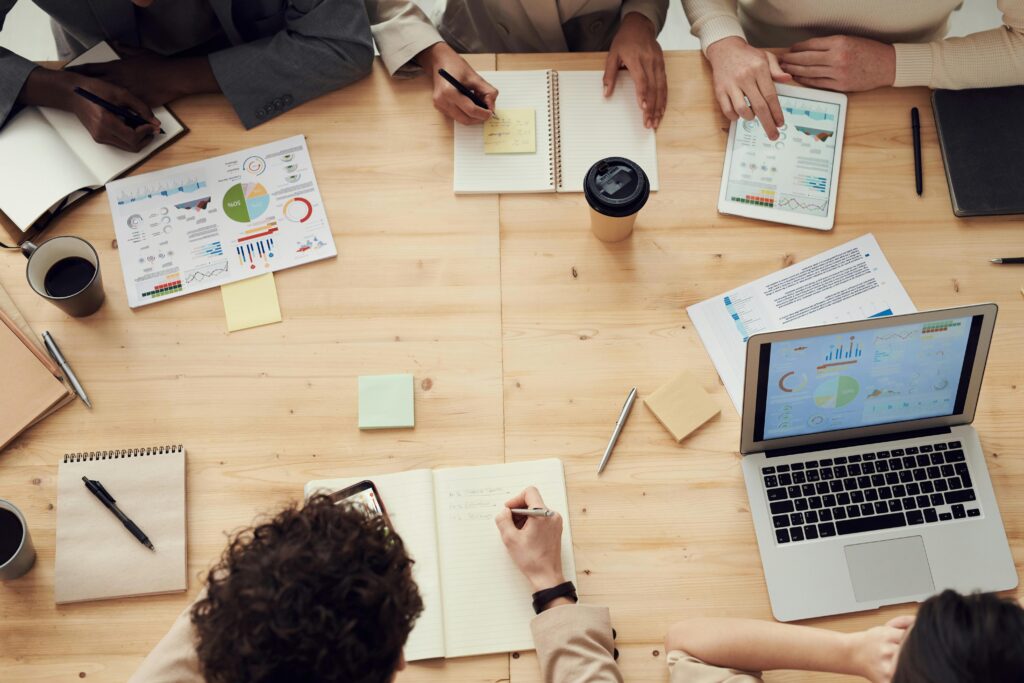 Overhead view of a group of people collaborating at a wooden table with laptops, charts, notebooks, and coffee cups, all engaged in reviewing data and taking notes.