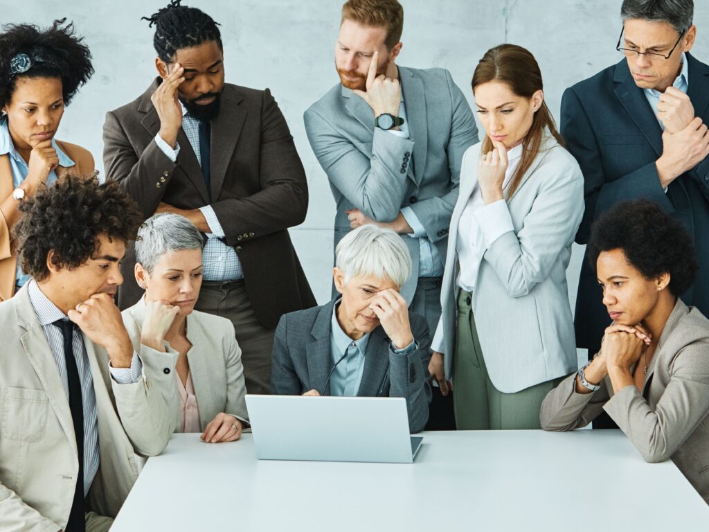 A group of business professionals, dressed in suits, are gathered around a laptop, looking concerned and deep in thought.